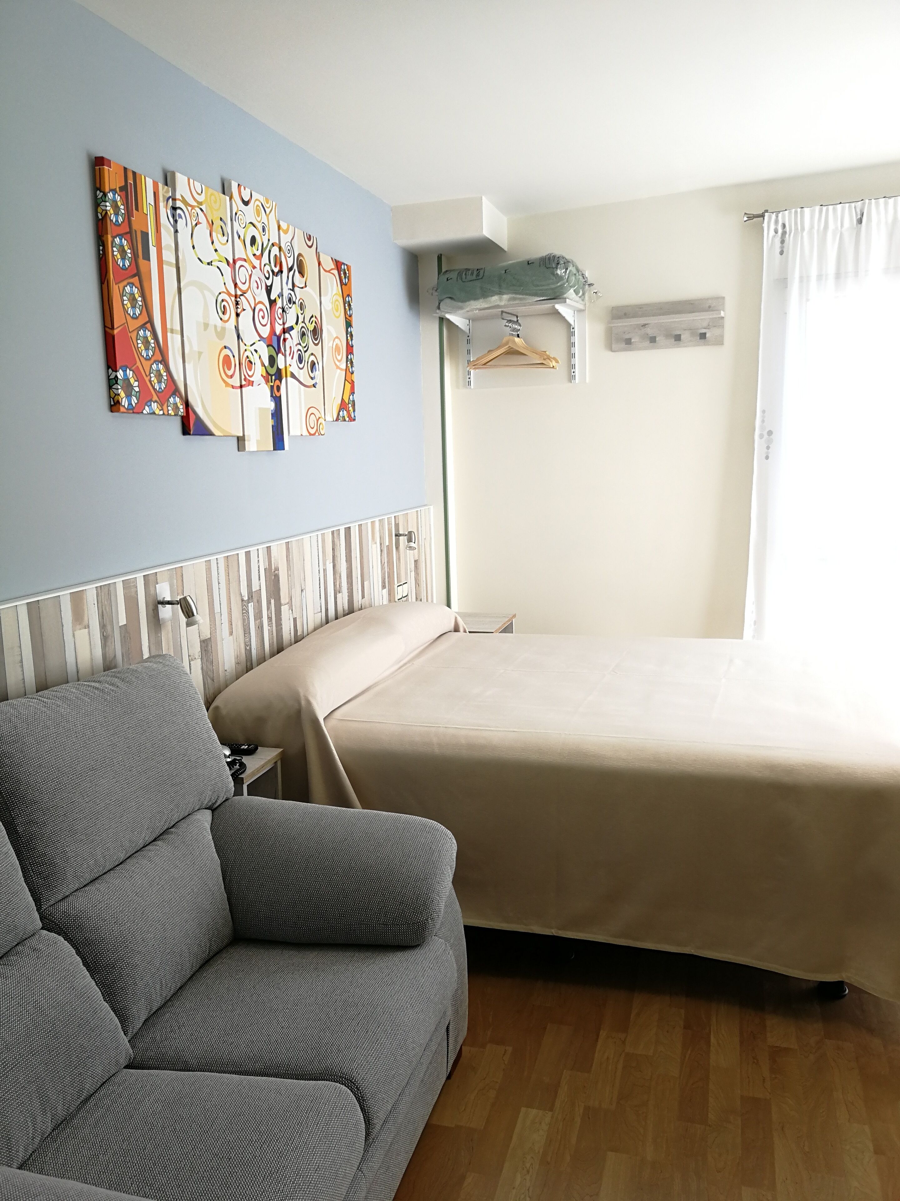 Habitació amb un llit, un sofà gris, un terra de fusta i una paret amb obra d'art colorida.