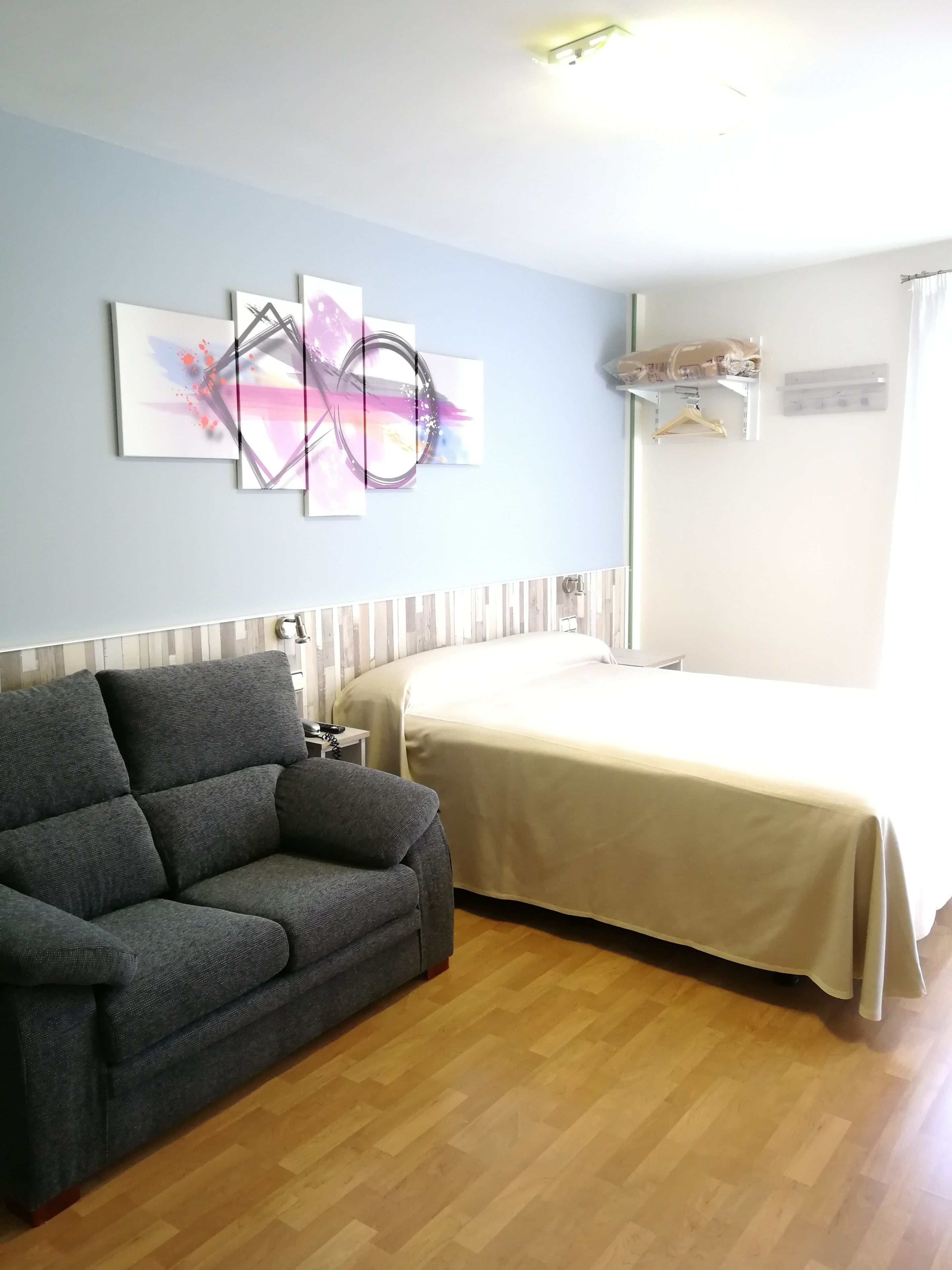 Una habitación con un sofá gris, una cama con cubrecama blanco y arte abstracto en la pared.
