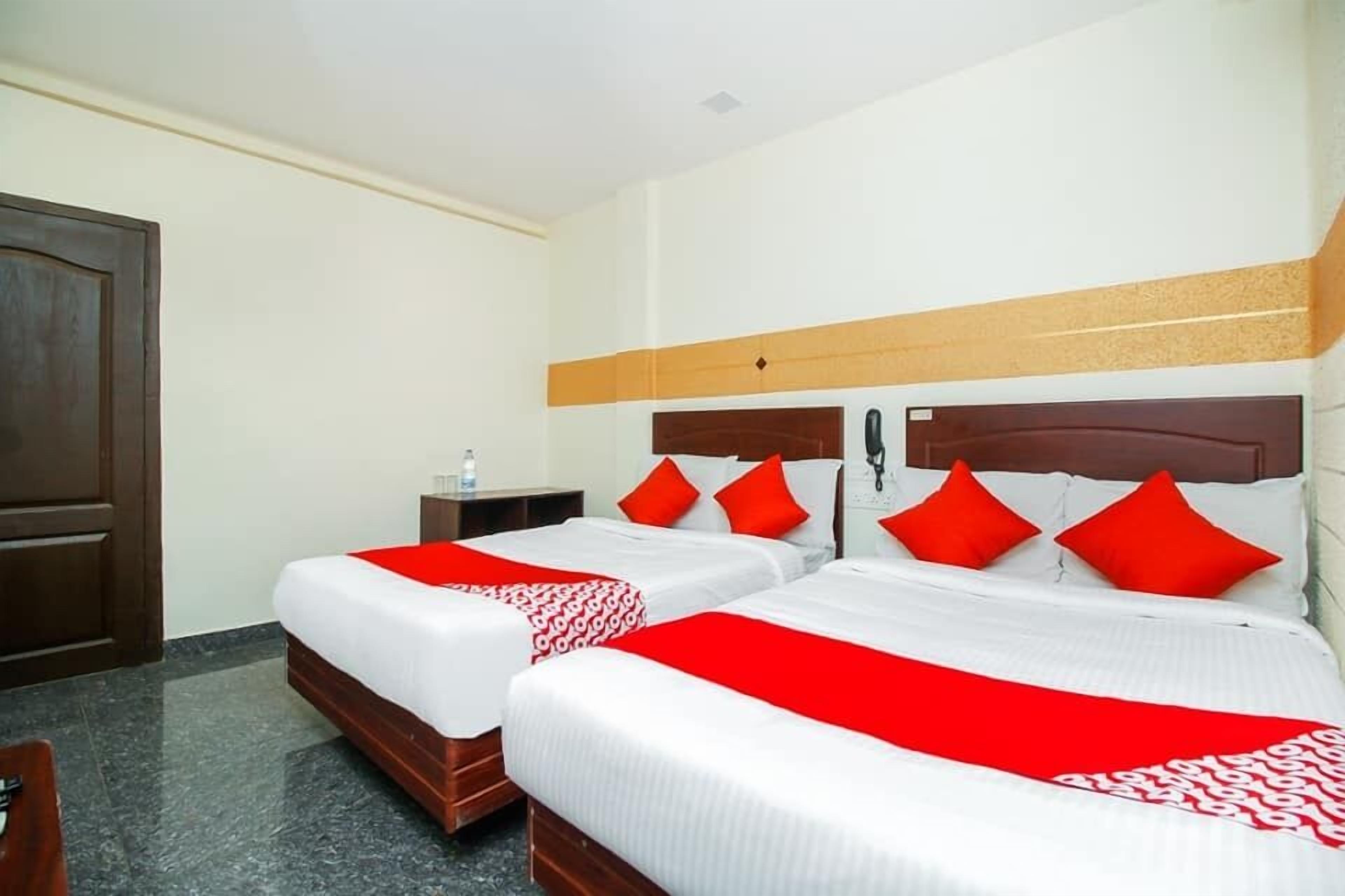 A hotel room with two beds, each with white bedding and red accents, a wooden headboard, a nightstand with a bottle, and a door.