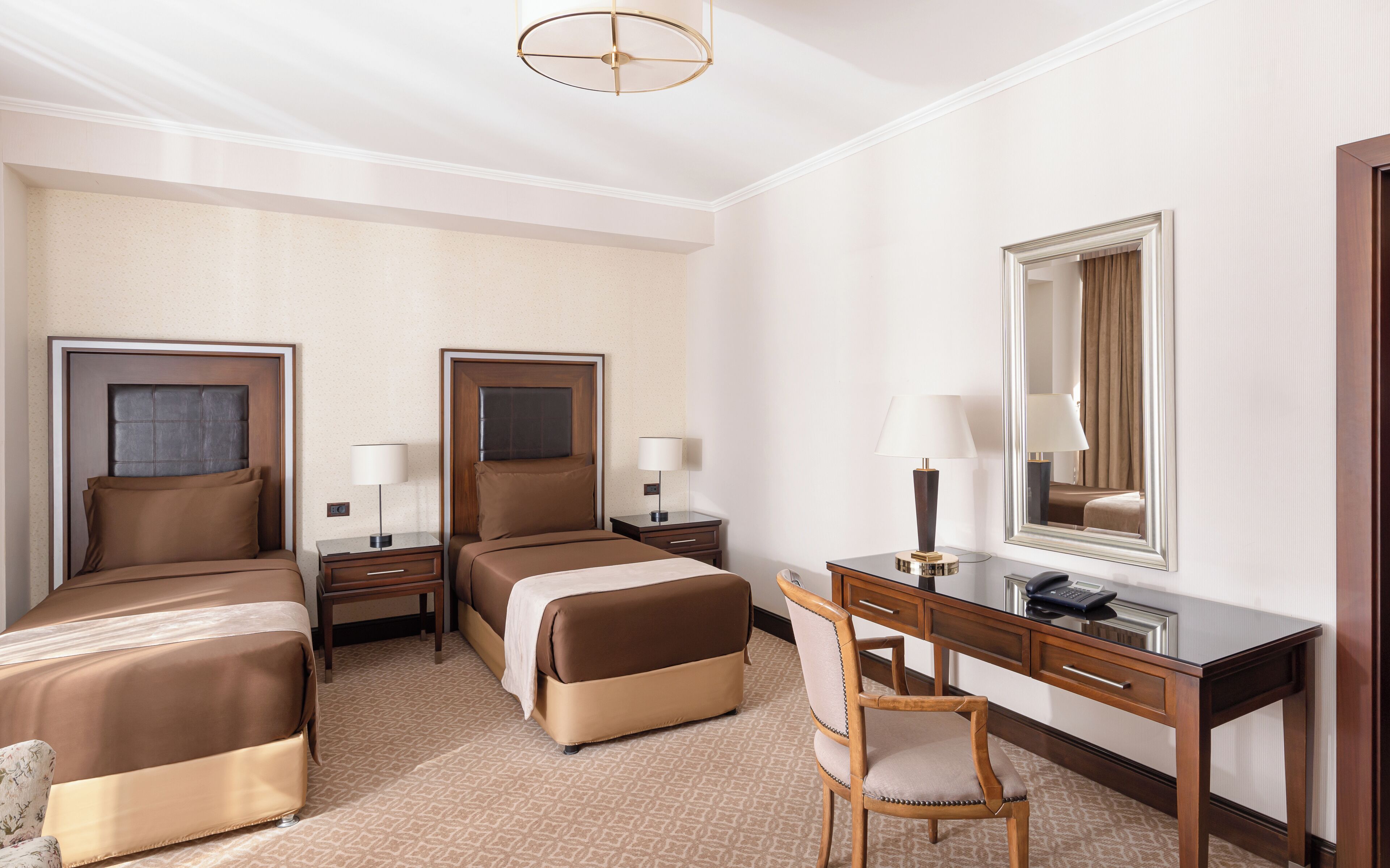A hotel room with two beds, a desk, a chair, a lamp, a telephone, and a mirror.