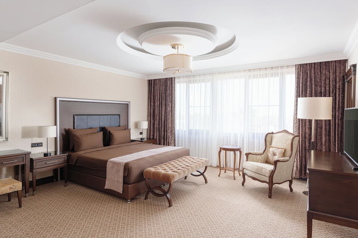 Quarto de hotel com uma cama grande, área de estar com uma cadeira e um banco, escrivaninha com luminária, televisão e luminária no teto.