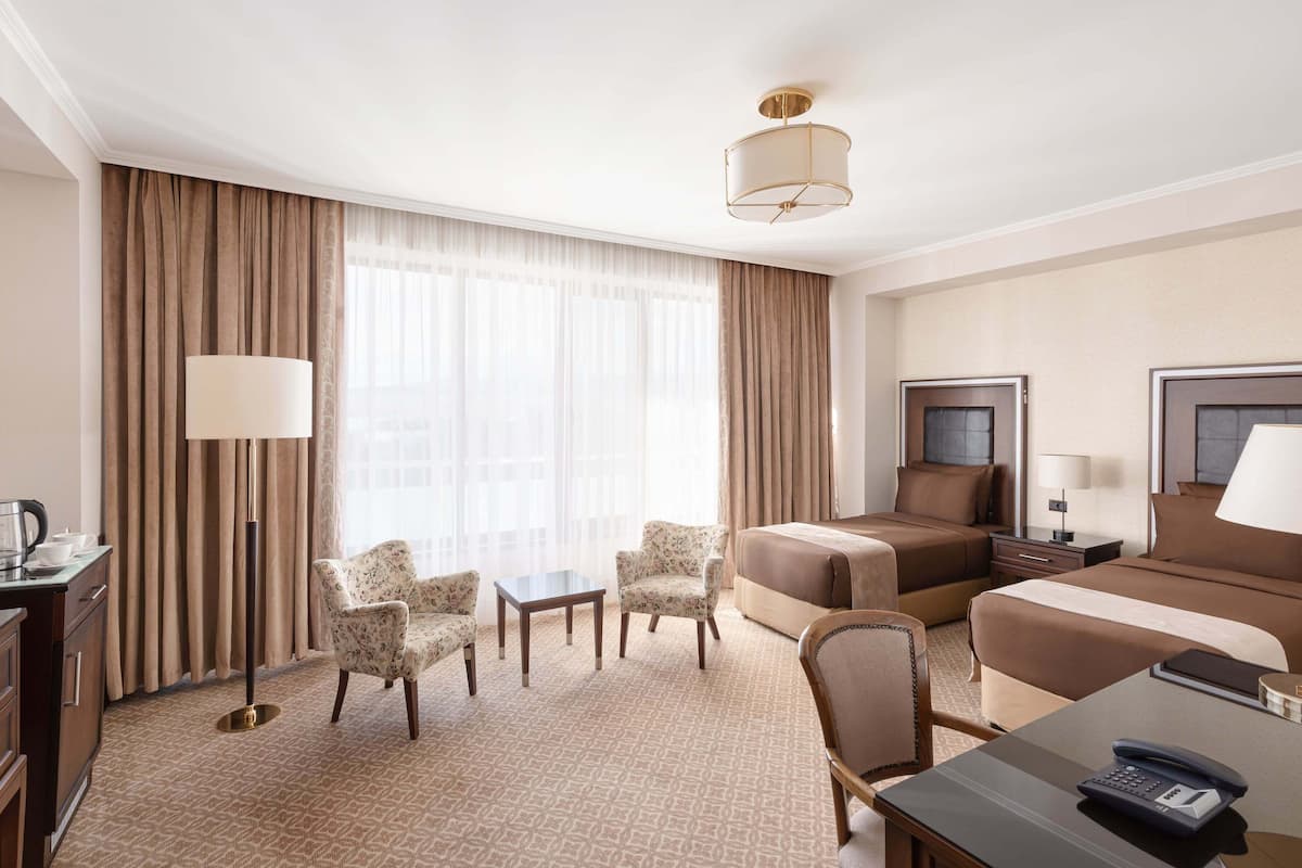 Quarto de hotel com cama, duas cadeiras, escrivaninha com telefone, luminária e uma grande janela com cortinas.