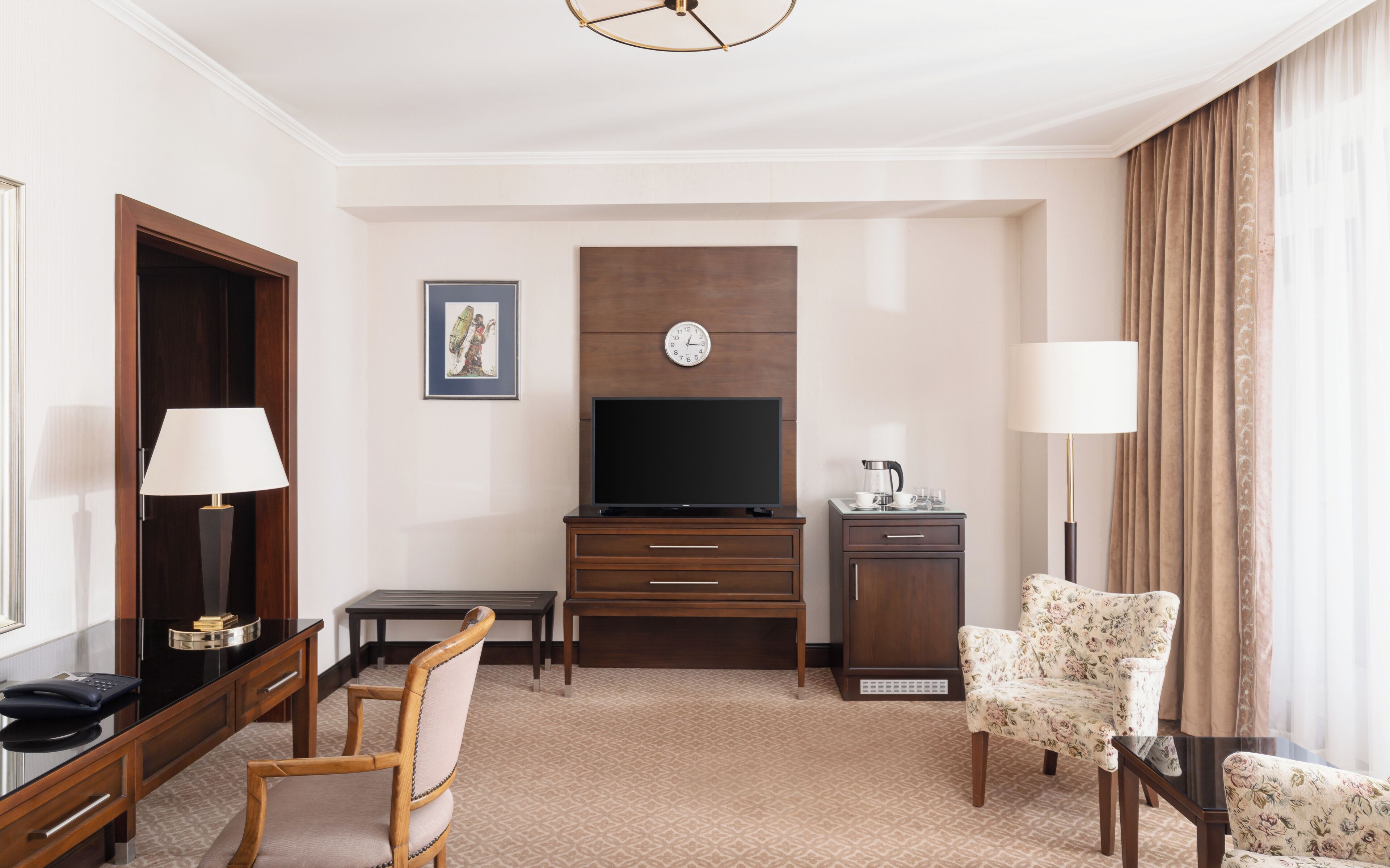 Een hotelkamer met een houten bureau, een stoel, een televisie, een klok, een lamp en een fauteuil met bloemenprint.
