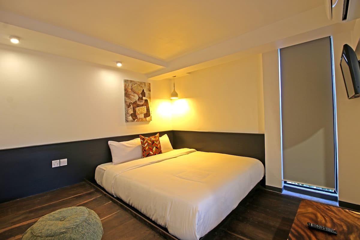 A neatly made bed with white bedding, a single pillow, and a decorative pillow on the headboard. There is a small wooden table with a remote control on it, and a wall-mounted TV to the right. The room has a dark wooden floor and a light-colored wall with a painting above the bed.