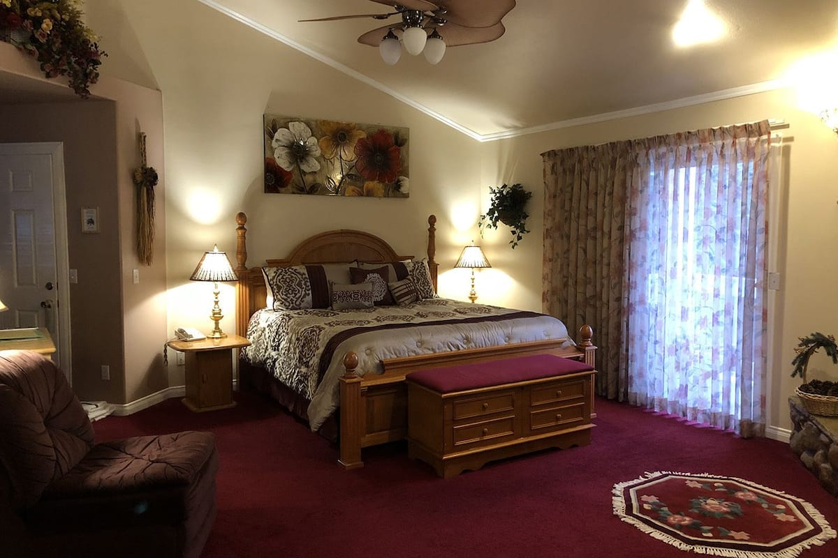 A bedroom with a large bed, two bedside tables with lamps, a wooden headboard, a ceiling fan, a floral painting, a window with curtains, and a red carpet.