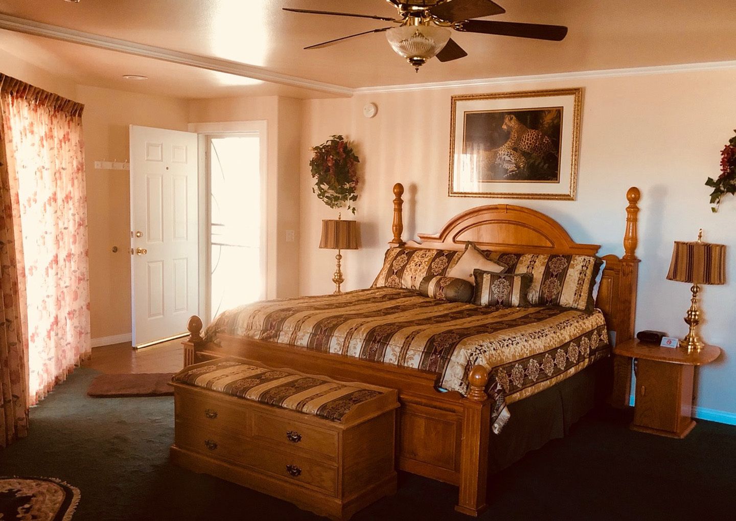 A bedroom with a wooden bed, a ceiling fan, a large window with curtains, a door, and a framed picture on the wall.