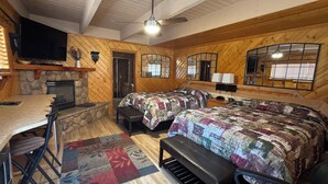 A cozy cabin with a stone fireplace, a large bed with a quilt, a dining area with a table and chairs, and a television mounted on the wall. - Giant Oaks Lodge (Running Springs)