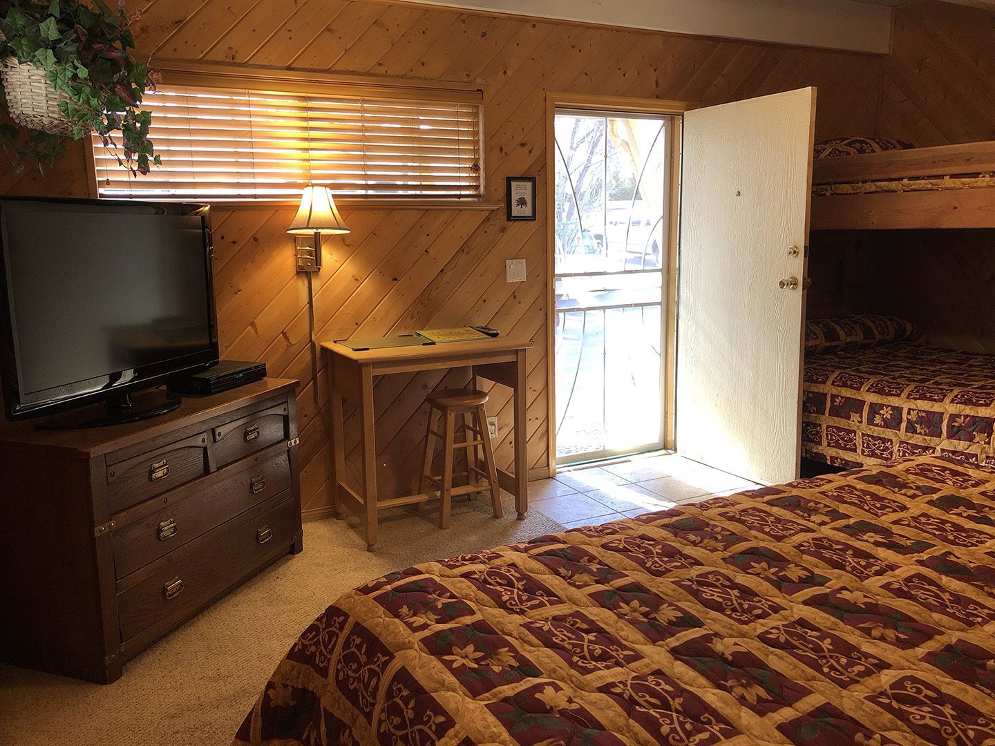 Une pièce comprenant une télévision posée sur une commode, un bureau avec une lampe, un lit recouvert d’une couvre-lit à motifs et une porte donnant sur un espace extérieur.