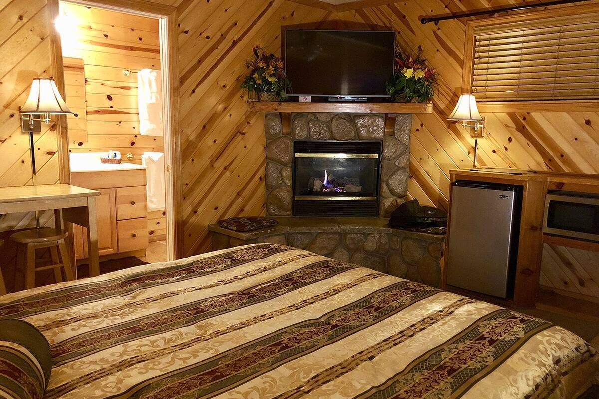 A cozy cabin-style room with a stone fireplace, a flat-screen TV mounted above it, a bed with patterned bedding, a small kitchenette, and a wooden desk with a lamp.