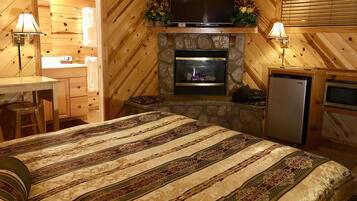 A cozy cabin-style room with a stone fireplace, a flat-screen TV mounted above it, a bed with patterned bedding, a small kitchenette, and a wooden desk with a lamp.