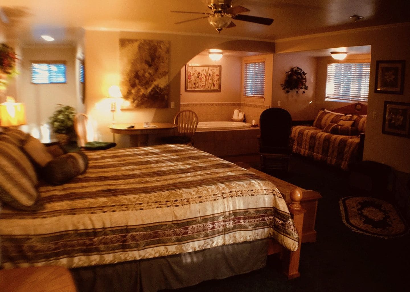 A bedroom with a large bed, a desk, a chair, a window with blinds, and a ceiling fan.
