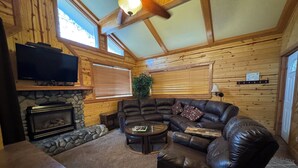 A cozy log cabin interior with a stone fireplace, leather sofa, and a television mounted above it. - Giant Oaks Lodge (Running Springs)