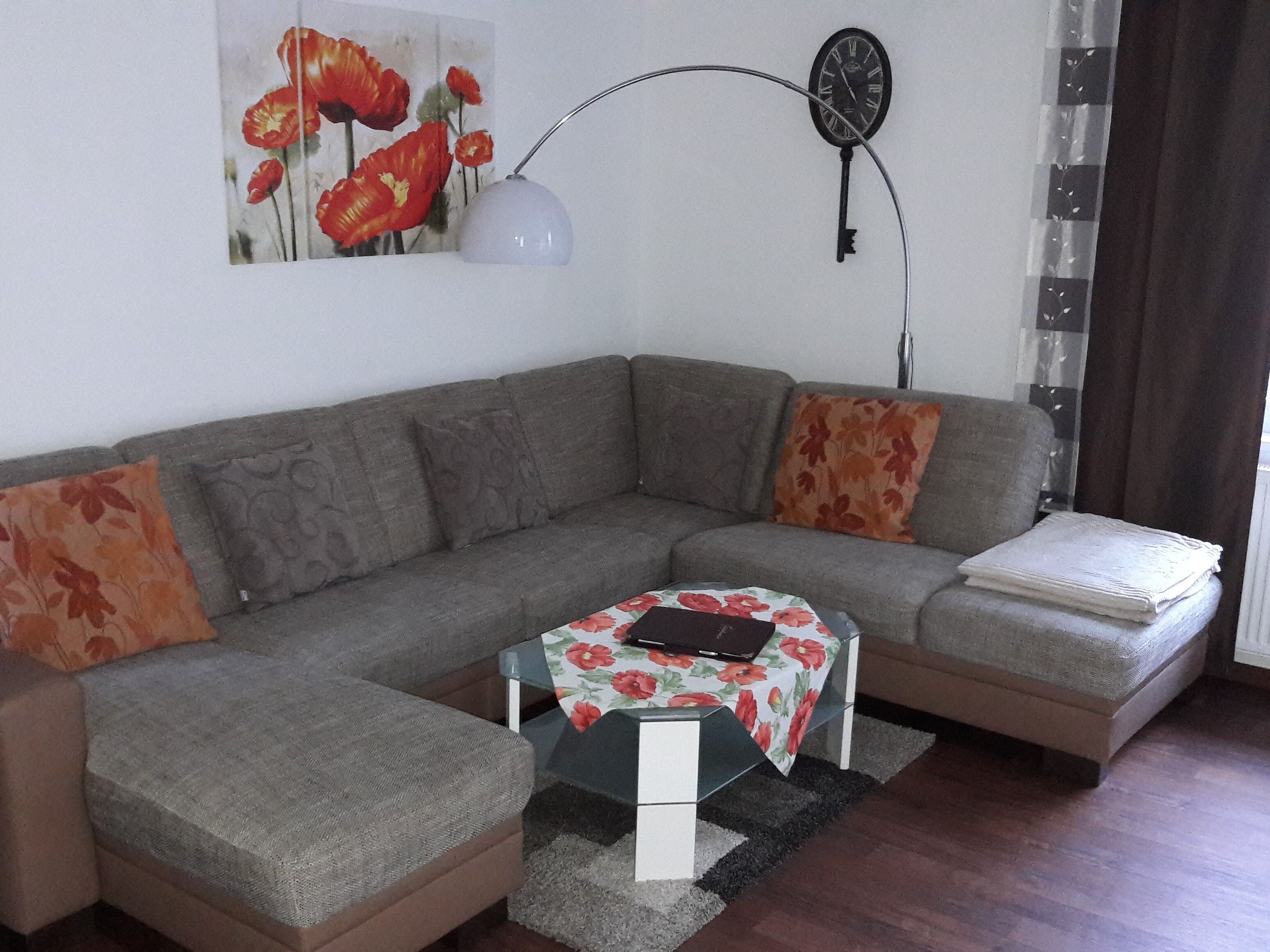 Un salon moderne avec un canapé d’angle gris, une petite table recouverte d’une nappe à motifs floraux et une horloge fixée au mur.