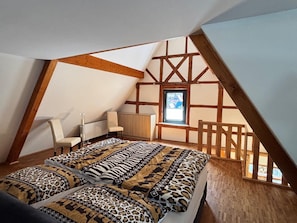 A loft bedroom with a sloped ceiling, exposed wooden beams, a bed with patterned bedding, a small window, and a chair.