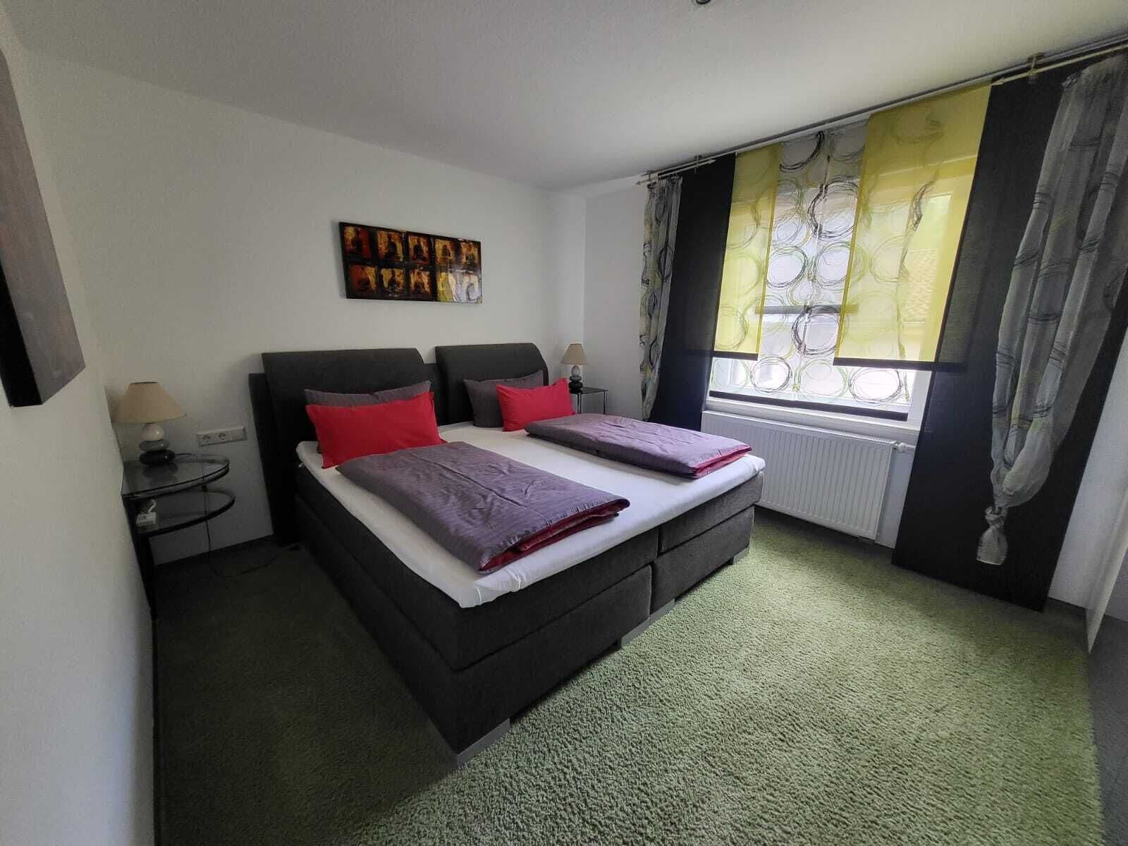 A double bed with red and purple bedding, a small bedside table with a lamp, and a window with patterned curtains.