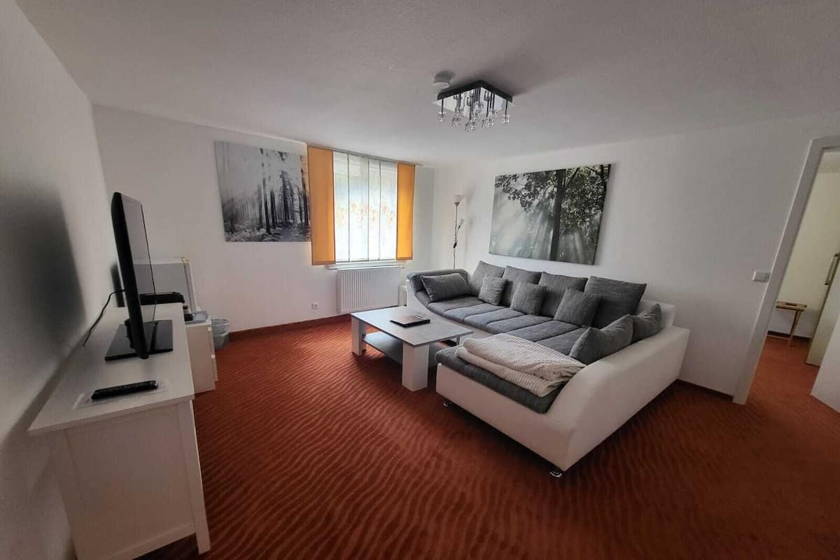 A modern living room with a grey sofa, a coffee table, and a TV on a white stand. There is a window with curtains, a painting on the wall, and a chandelier.