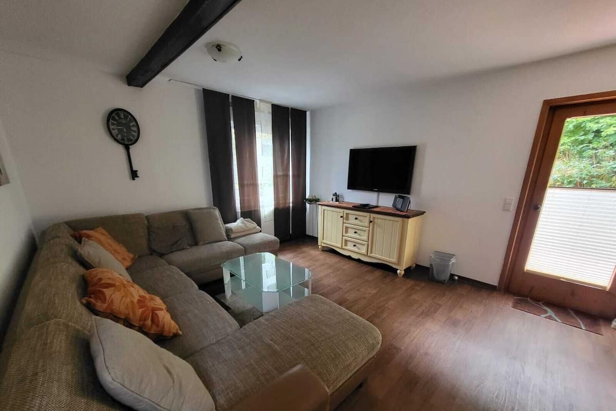 A living room with a sofa, a glass coffee table, a television on a cabinet, and a door leading outside.