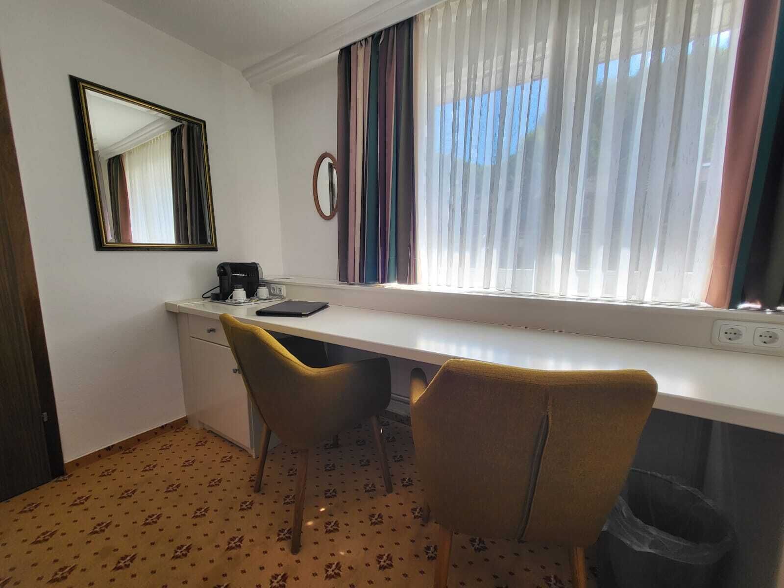 A hotel room with a white desk, two chairs, a mirror, a window with curtains, and a patterned carpet.