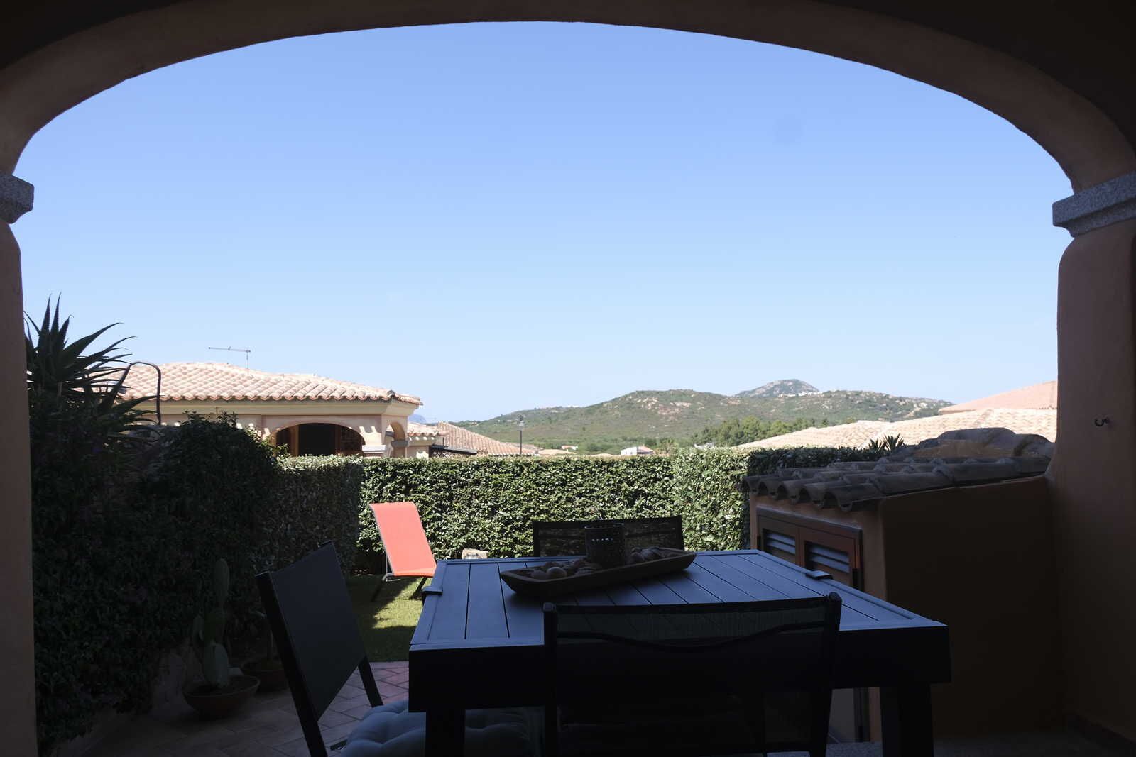Une terrasse extérieure avec une table et des chaises, une vue sur un bâtiment avec un toit en tuiles et un paysage montagneux en arrière-plan.