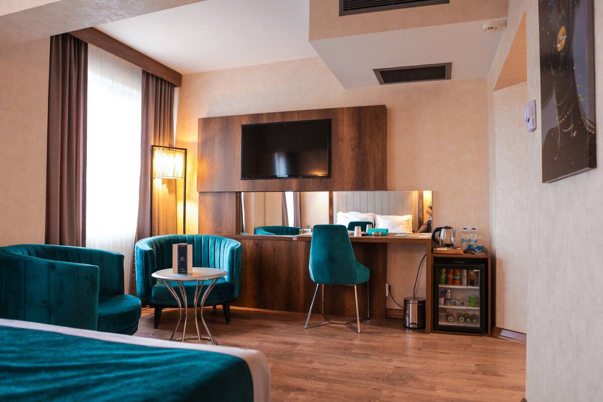 Una habitación de hotel con una cama, dos sillones turquesa, una mesita redonda pequeña, un escritorio con una silla, un televisor montado en la pared y un minibar.
