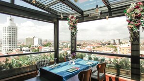 Un coin repas avec une table dressée pour quatre personnes, de grandes fenêtres donnant sur la ville et des compositions florales suspendues.