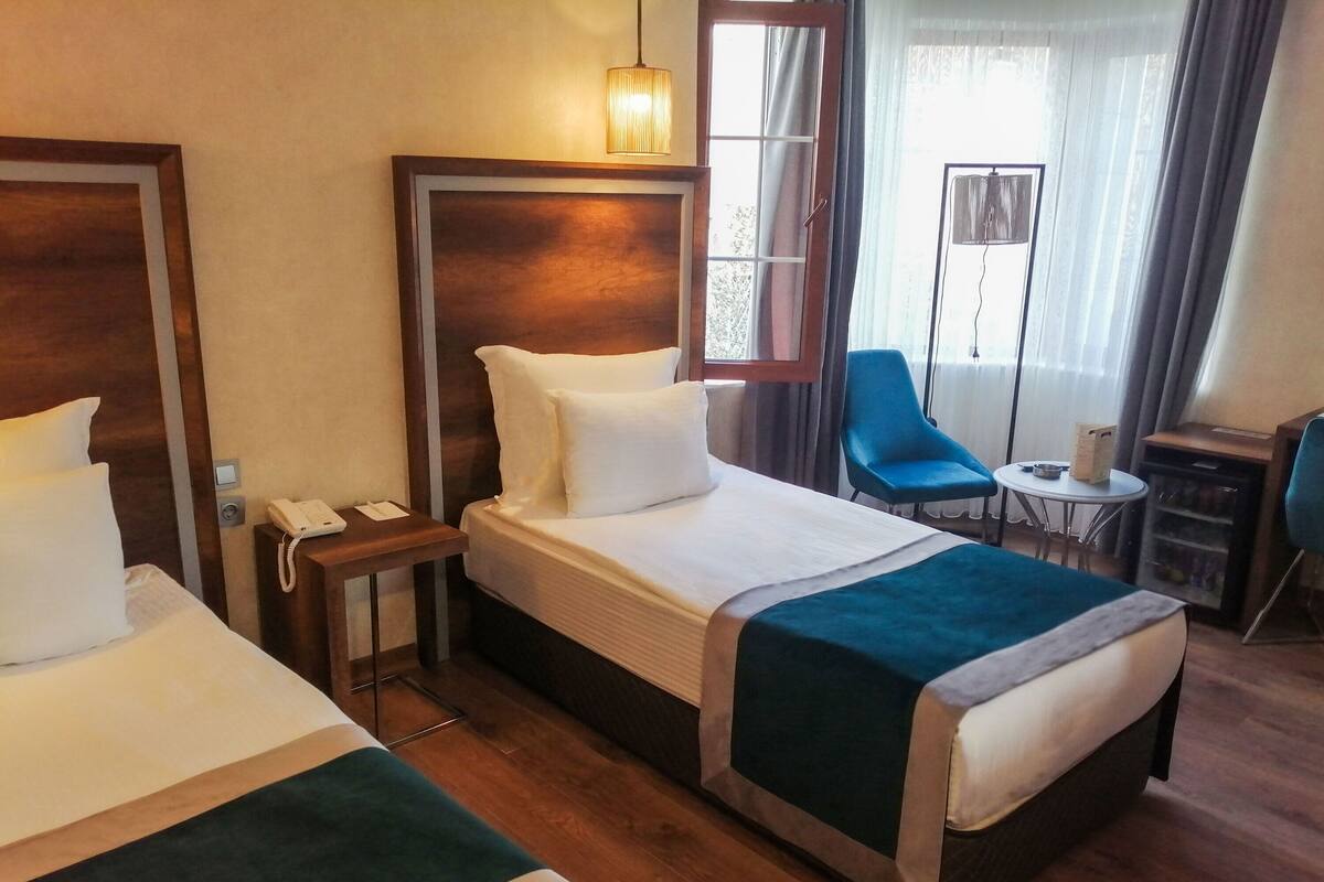 Habitación de hotel con dos camas, un escritorio con teléfono, una silla, una mesita y una ventana con cortinas.