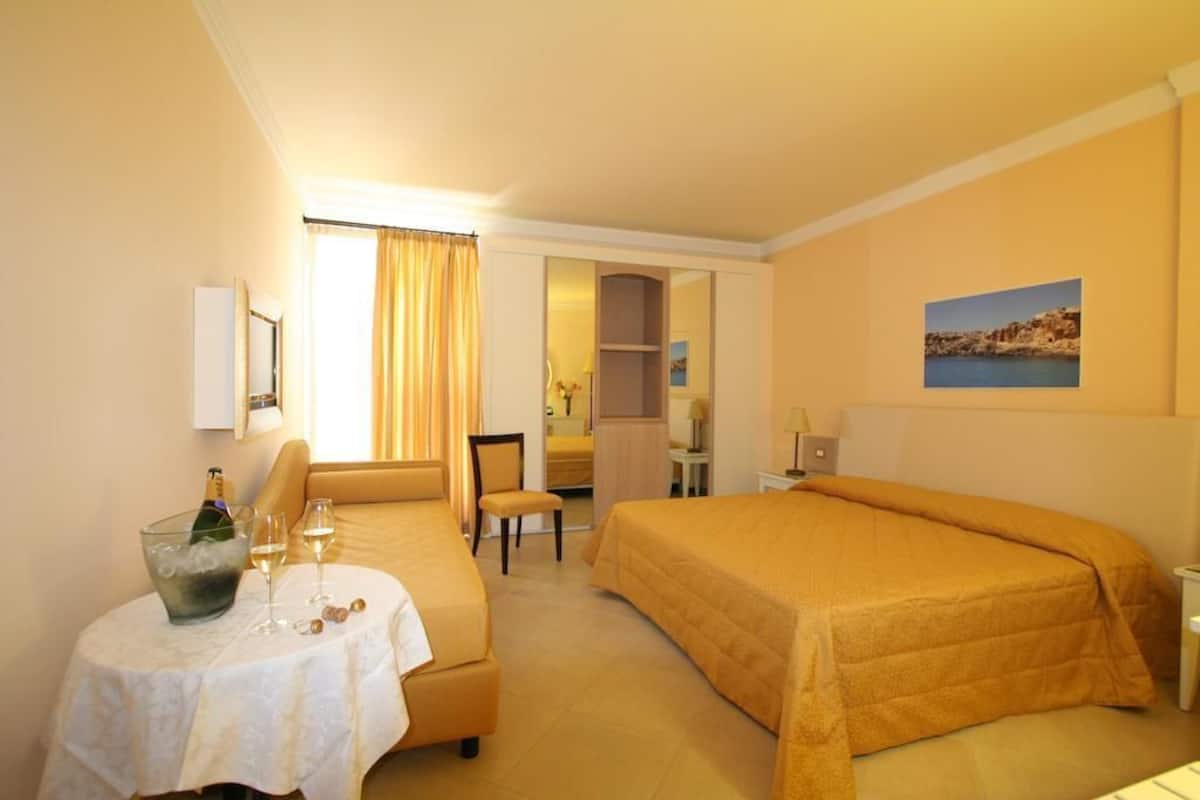 A hotel room with a bed, a small table with a bottle and glasses, a chair, and a desk.