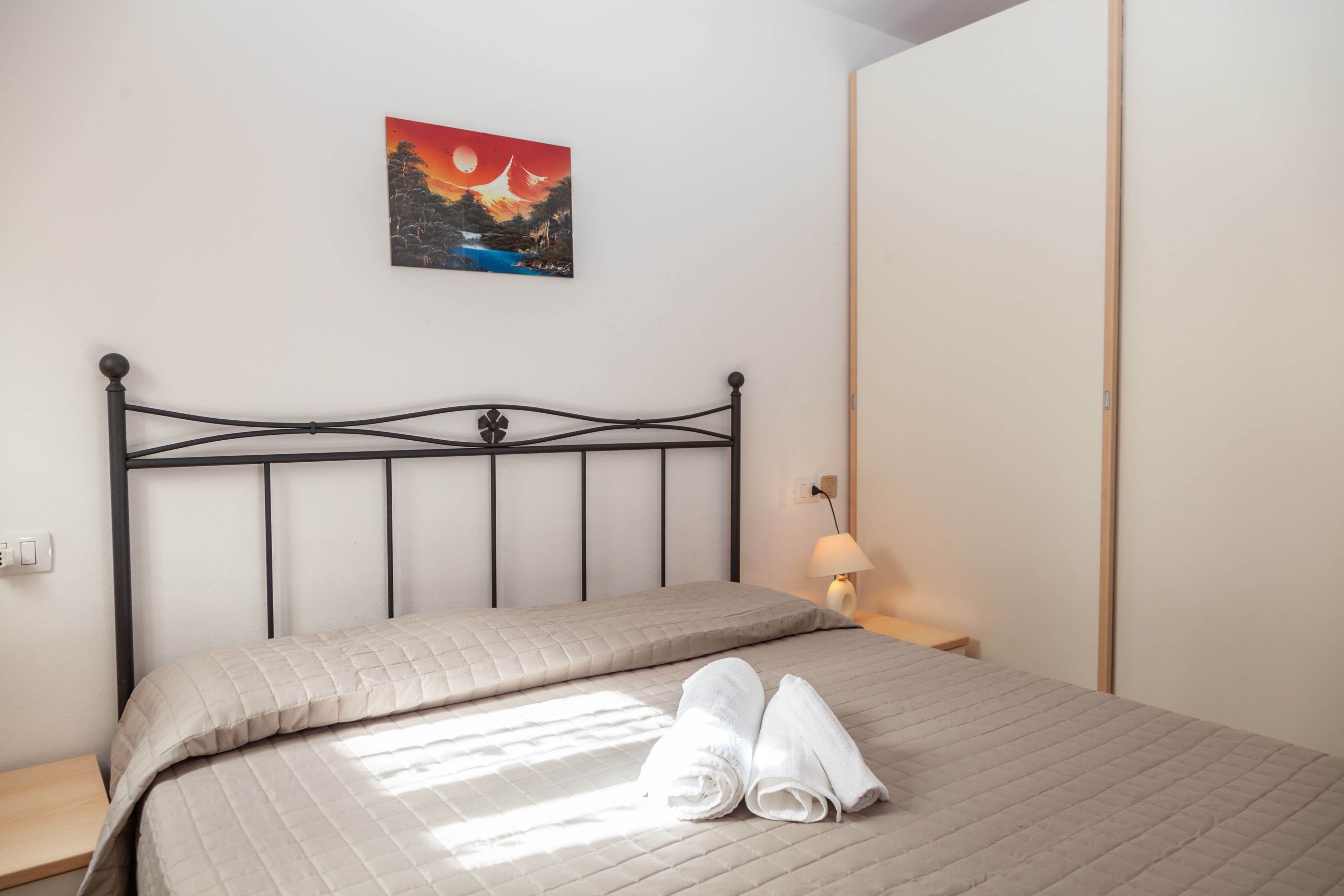 Un lit bien fait, recouvert d’une courtepointe matelassée, avec deux serviettes enroulées, un tableau représentant un coucher de soleil accroché au mur et une lampe de chevet posée sur une table de nuit.