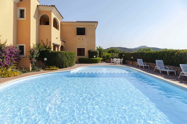 Uma piscina com espreguiçadeiras, um edifício com arcos e um cenário montanhoso ao fundo.