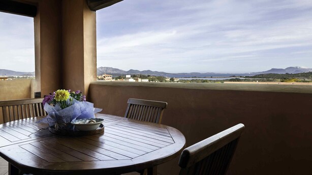 A wooden table with a floral centerpiece on a balcony with a view of distant mountains and buildings. - Aria di vacanza - Borgo Turchese (Olbia)