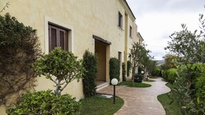 A pathway leading to a building with a yellow facade, surrounded by greenery and a paved walkway. - Aria di vacanza - Borgo Turchese (Olbia)
