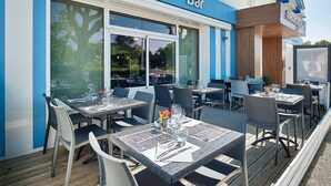 Uma área externa de restaurante com mesas e cadeiras, vista desobstruída para a rua e um letreiro com a inscrição "Restaurant - Bar".