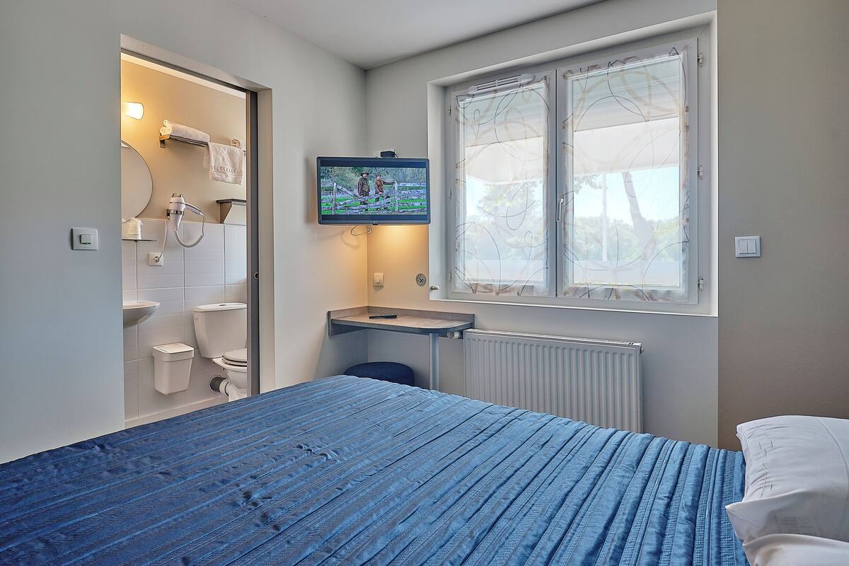 A bedroom with a bed, a TV mounted above a small table, a window with a view, and a bathroom with a toilet and sink.