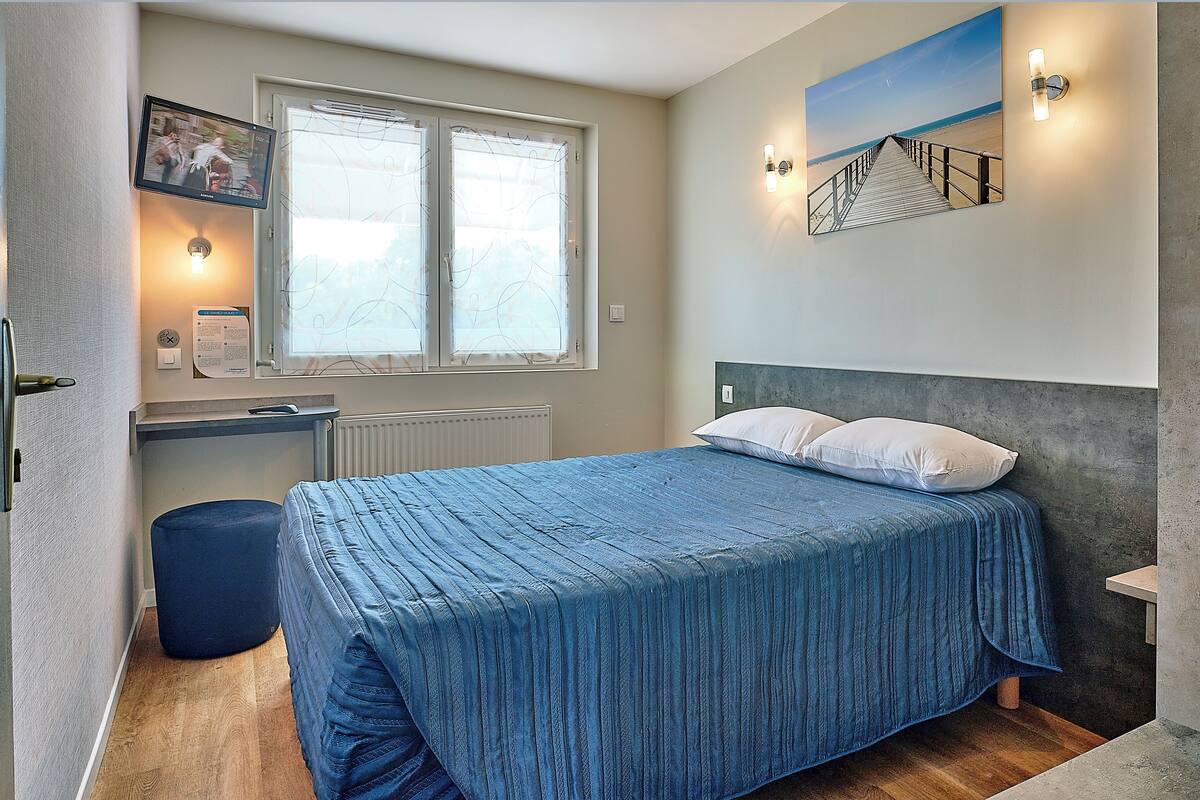 A bedroom with a bed, a window with blinds, a wall-mounted TV, and a painting of a pier.