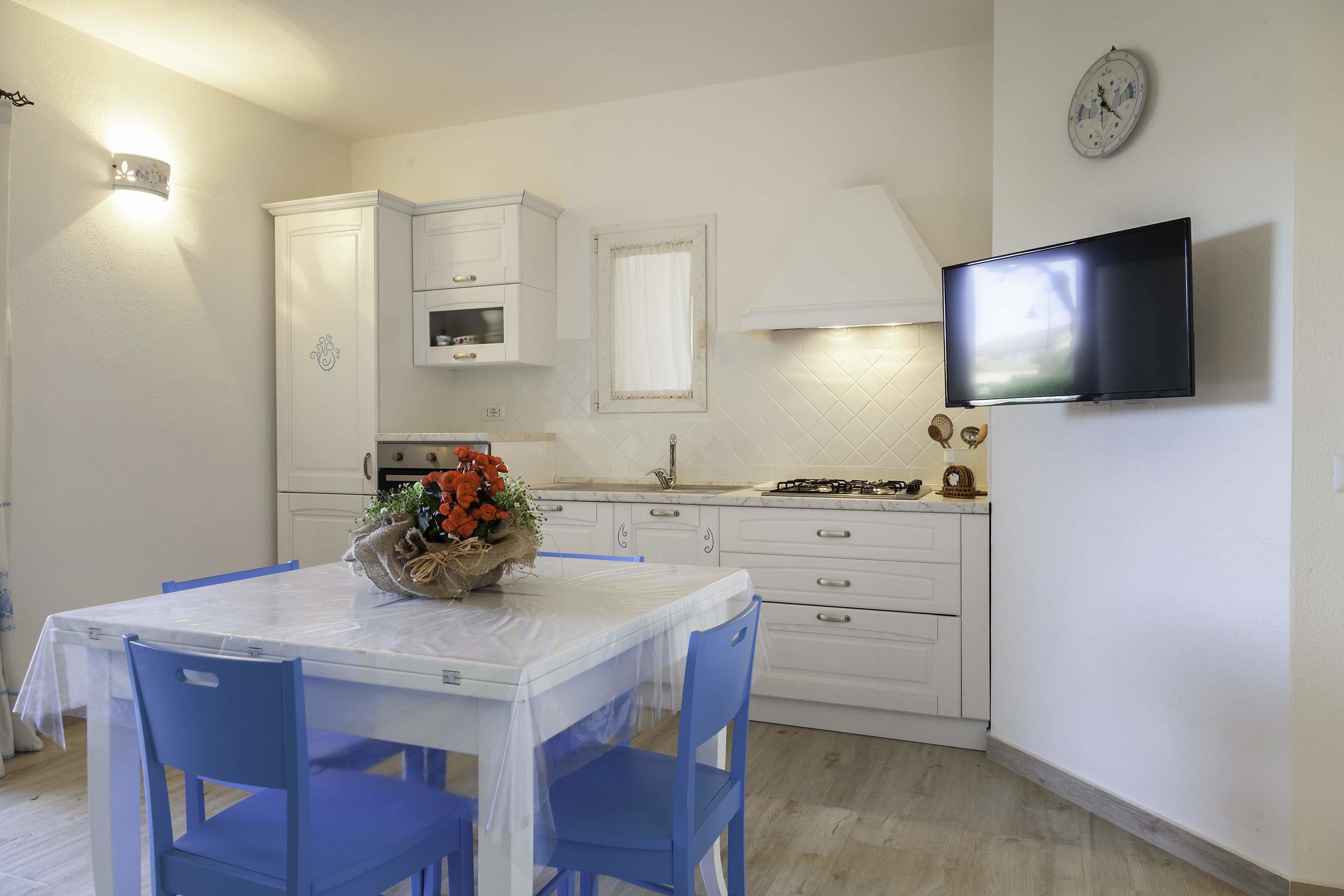 A modern kitchen with white cabinets, a dining table with blue chairs, a TV mounted on the wall, and a clock.