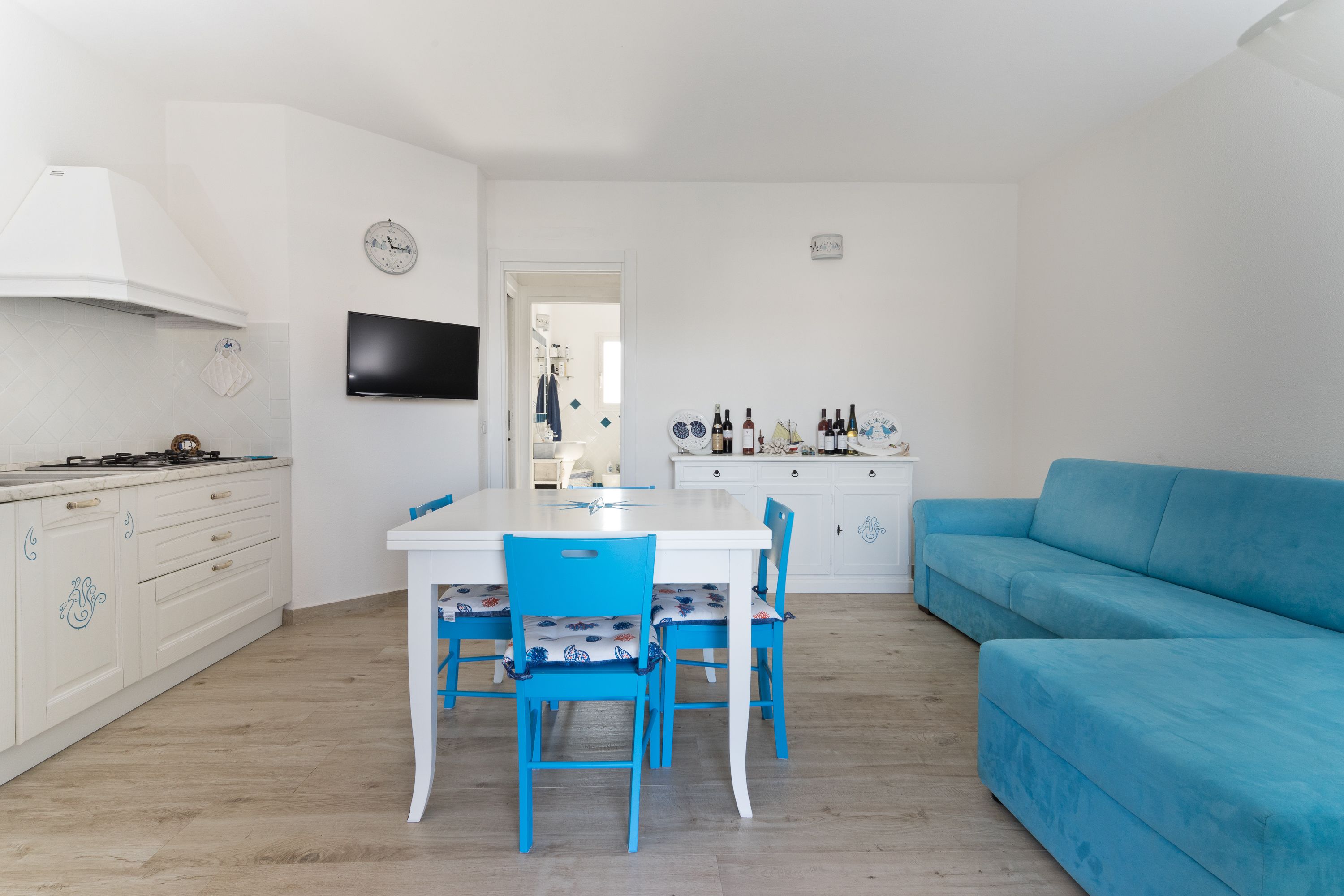 Uma cozinha compacta com mesa de jantar, cadeiras, um sofá azul e uma TV fixada na parede.