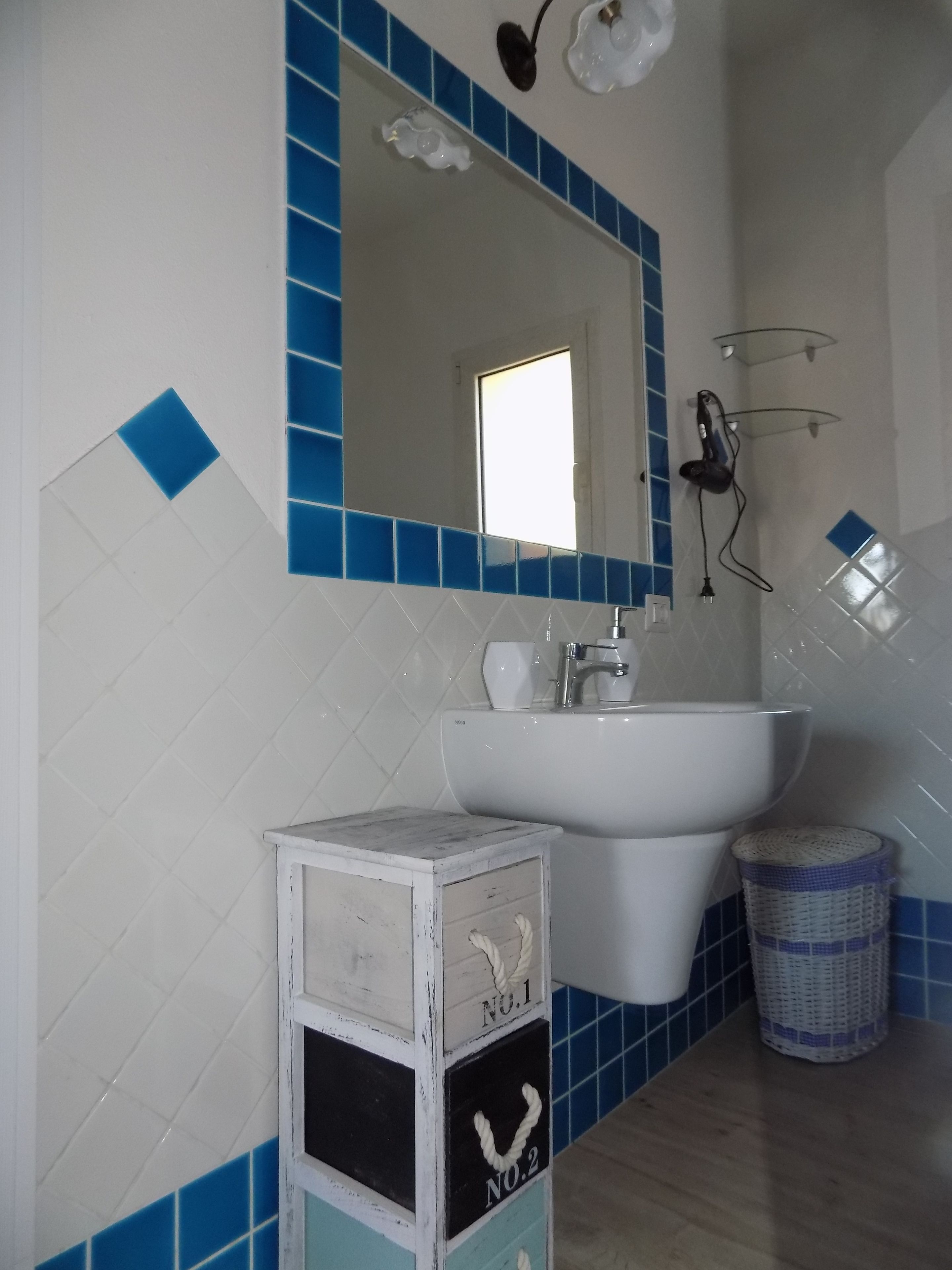 Een badkamer met een wit wastafel, blauwe betegelde wanden, een wit kastje met lades, en een rieten mandje.