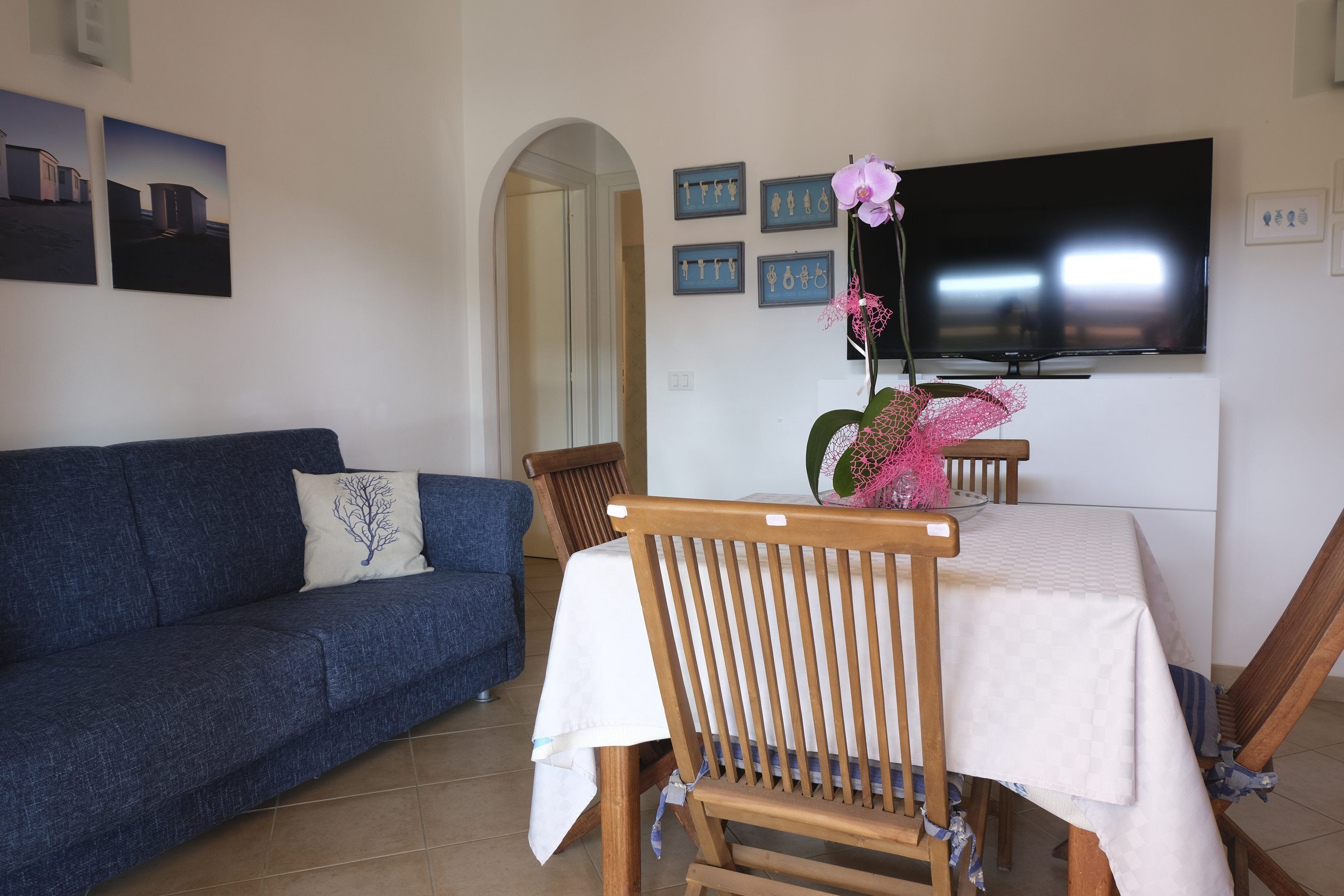 Ein Wohnzimmer mit einem blauen Sofa, einem Holzesstisch mit weißer Tischdecke und einem an der Wand befestigten Fernseher.