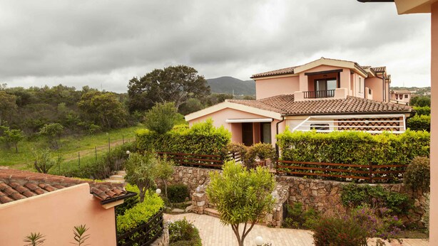 A two-story house with a tiled roof, surrounded by lush greenery and a stone wall. - Aria di Vacanza - S'Abba Lughente (Olbia)