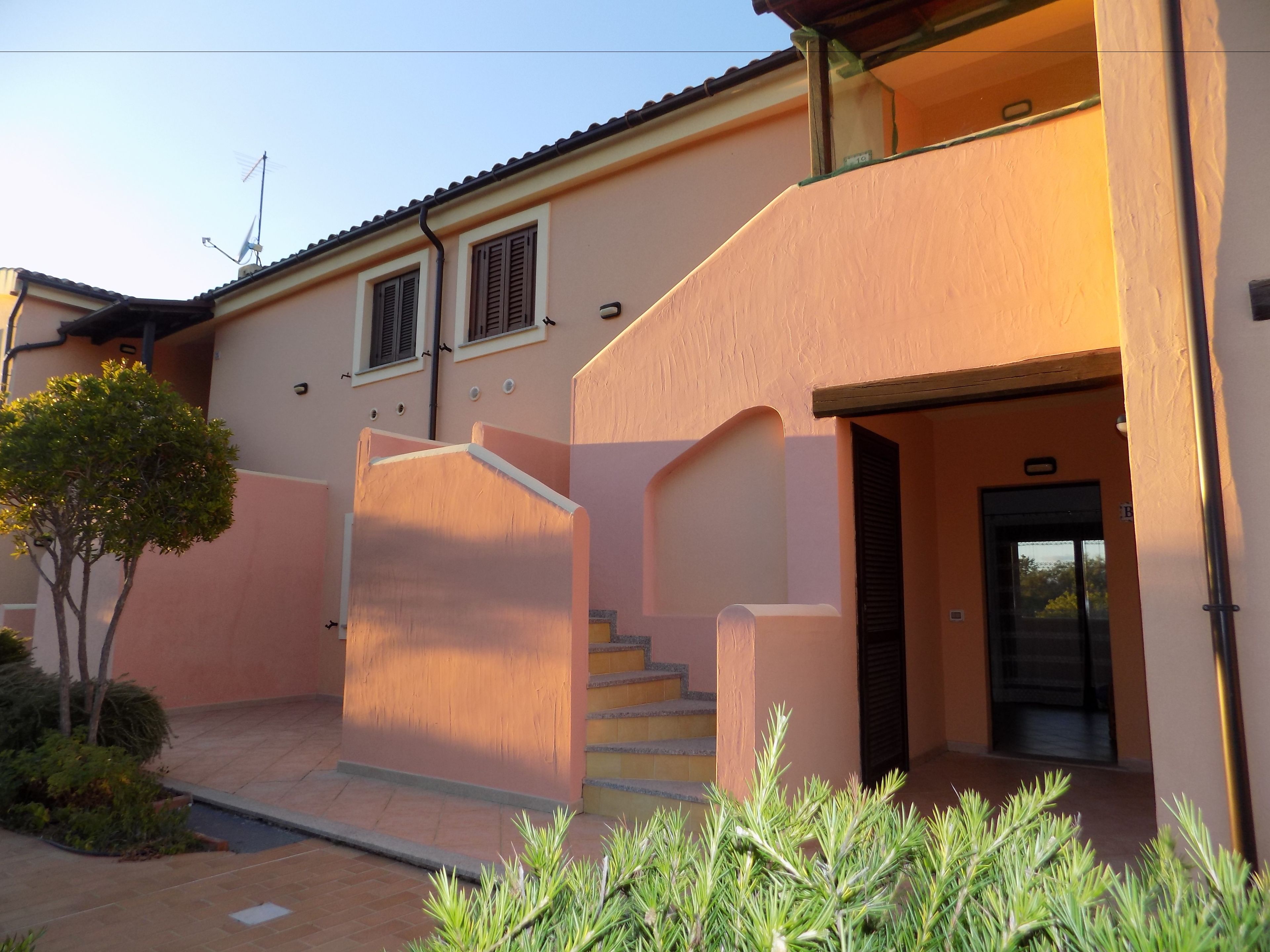 Un edificio de dos pisos con fachada color crema, una escalera que conduce a la entrada y un pequeño árbol en primer plano.