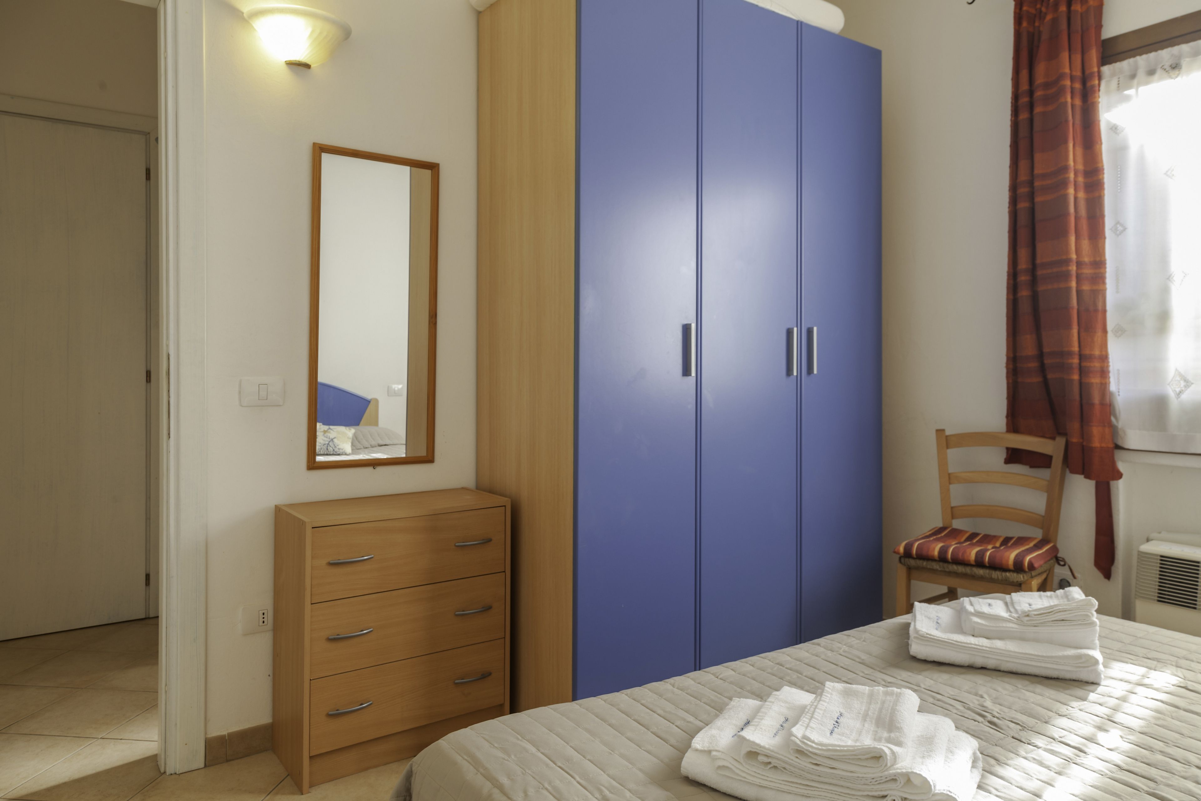 A bedroom with a bed, a wooden dresser, a blue wardrobe, a chair, and a mirror.