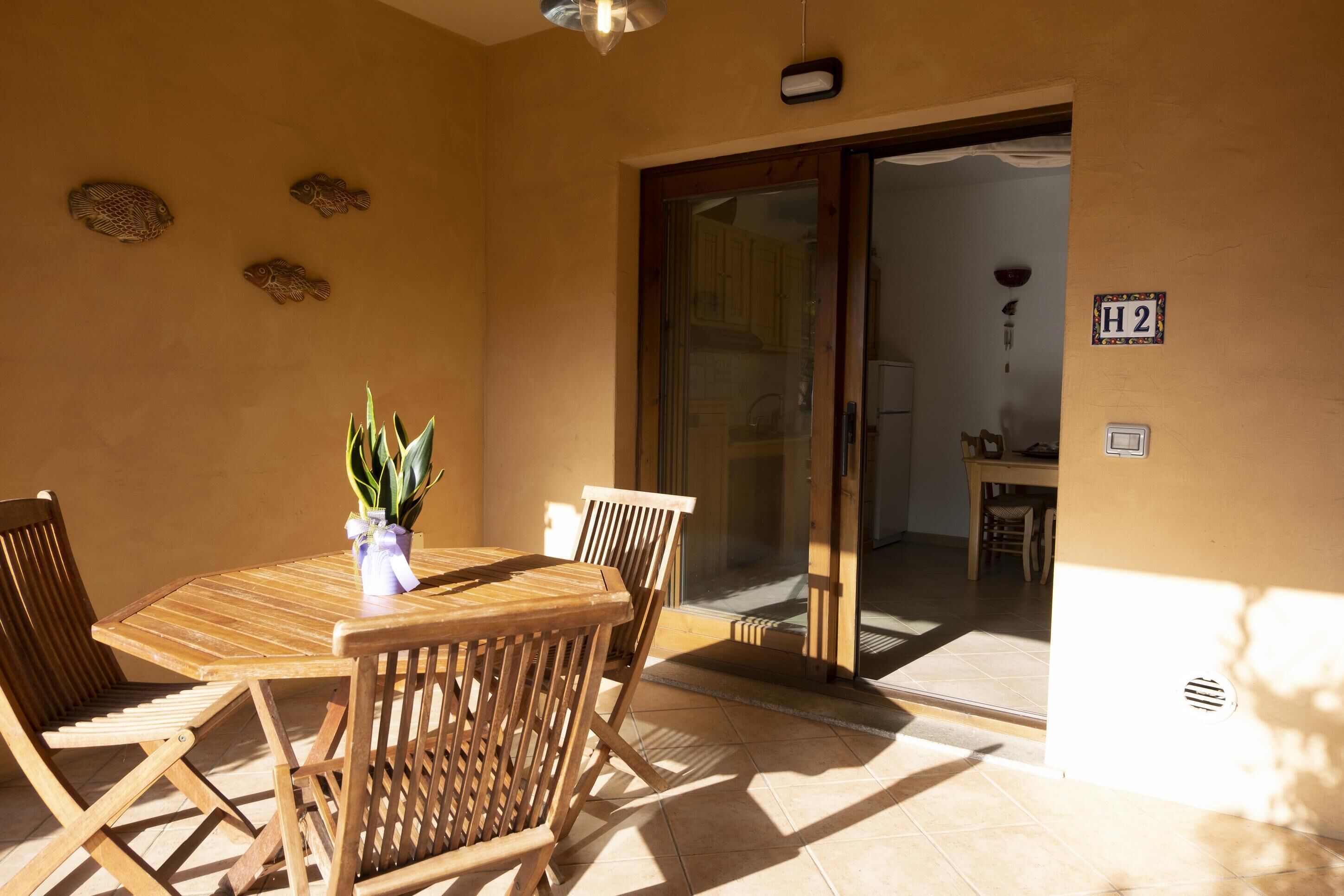 A sunlit patio with a wooden table and chairs, a potted plant, and decorative fish wall art.