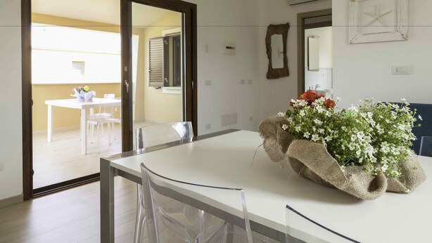 Un comedor con una mesa y sillas, un jarrón de flores y vista a otra habitación con una mesa y sillas.