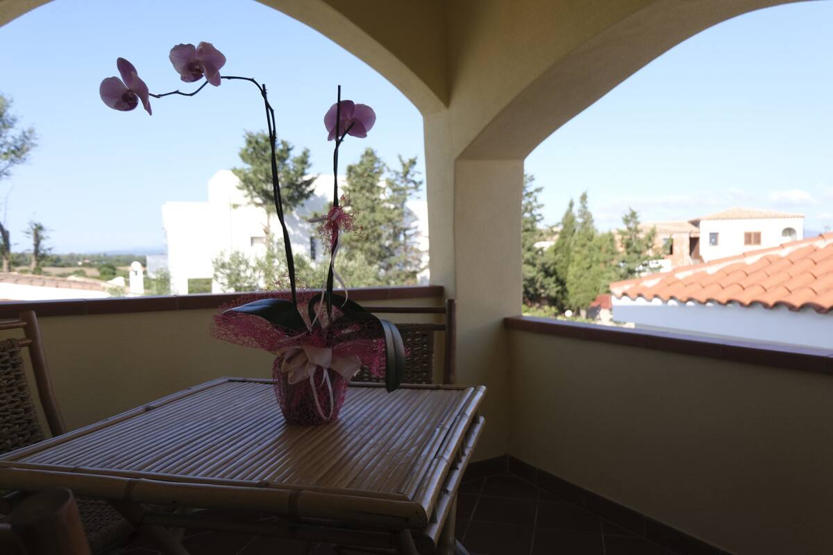 A balcony with a table and chairs, a vase with flowers, and a view of buildings and trees.