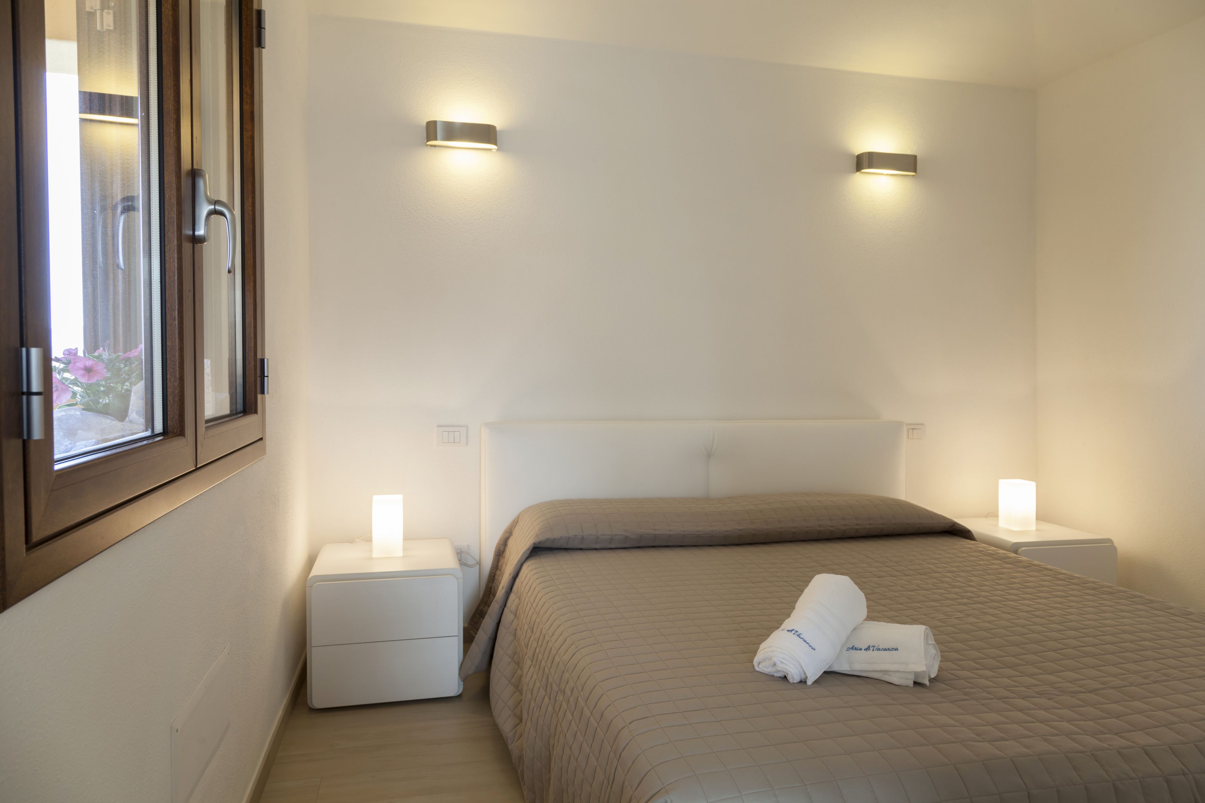 A hotel room with a bed, two bedside tables, a window, and wall-mounted lights.