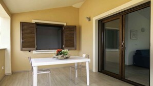 Un pequeño comedor con una mesa y sillas blancas, una ventana con contraventanas de madera y un portón corredera de vidrio que da a un balcón.