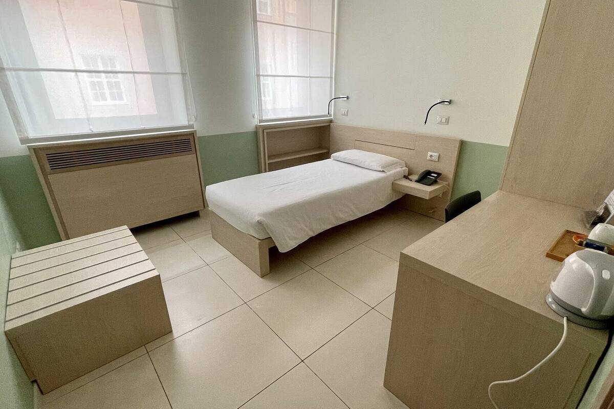Ein kleines, kompaktes Hotelzimmer mit einem Einzelbett, einem Nachttisch, einem kleinen Schreibtisch mit Telefon und einem Heizkörper.