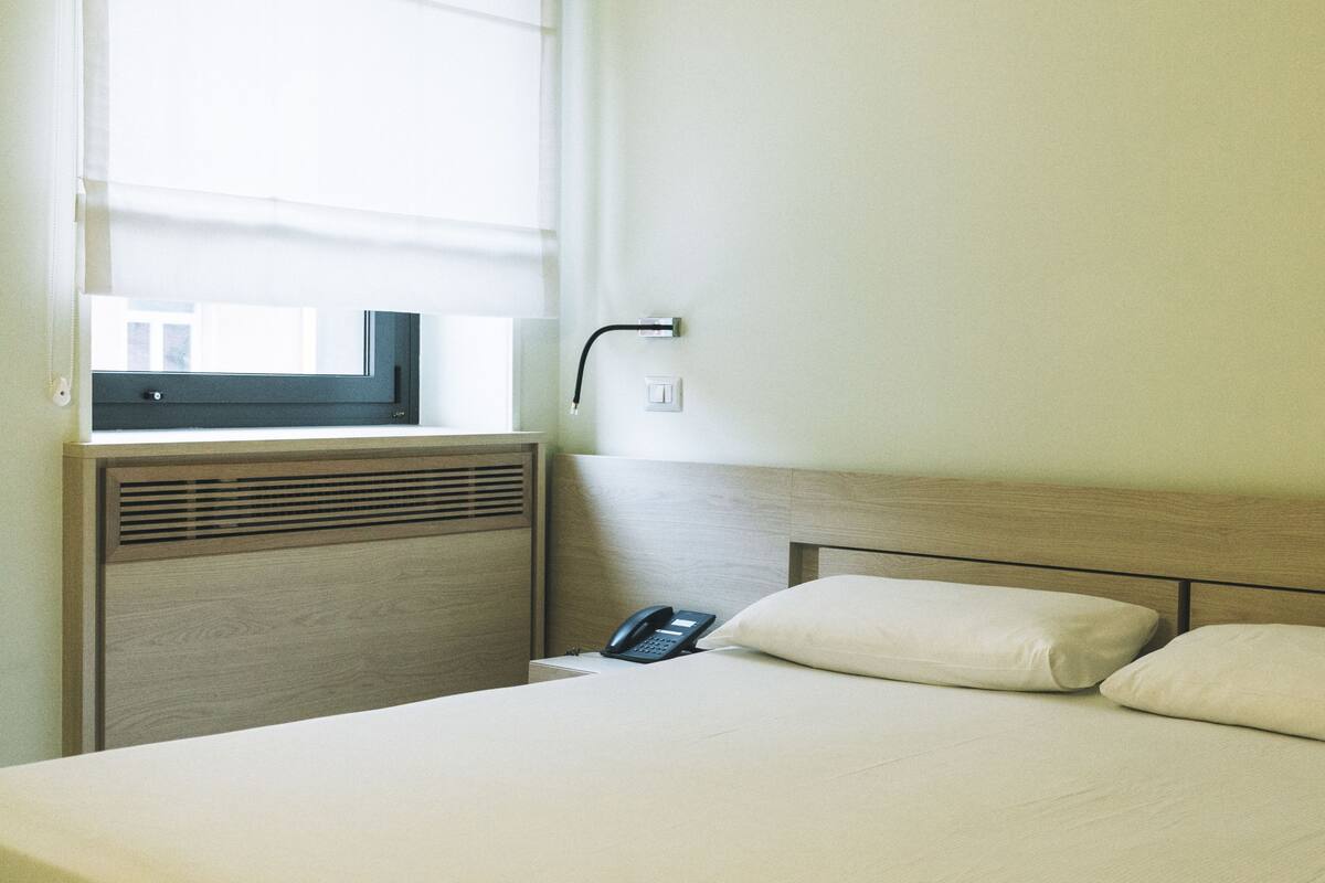 Ein Hotelzimmer mit einem Bett, einem Nachttischtelefon, einem Fenster mit Jalousien und einem an der Wand montierten Schalter.