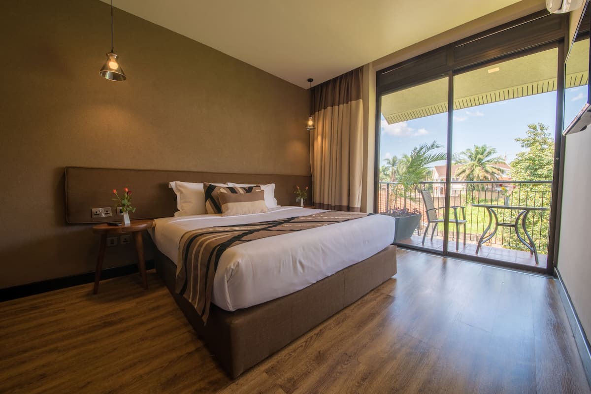 A spacious bedroom with a large bed, a nightstand, a potted plant, and a view of a balcony with outdoor furniture.