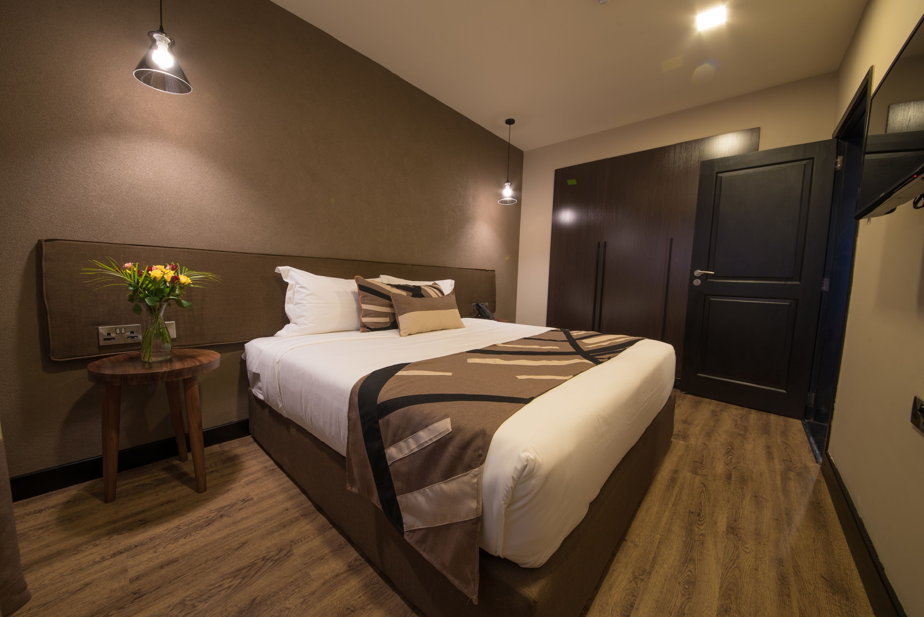 Une chambre d’hôtel avec un grand lit, une table de chevet ornée d’un vase de fleurs, une armoire et un téléviseur fixé au mur.